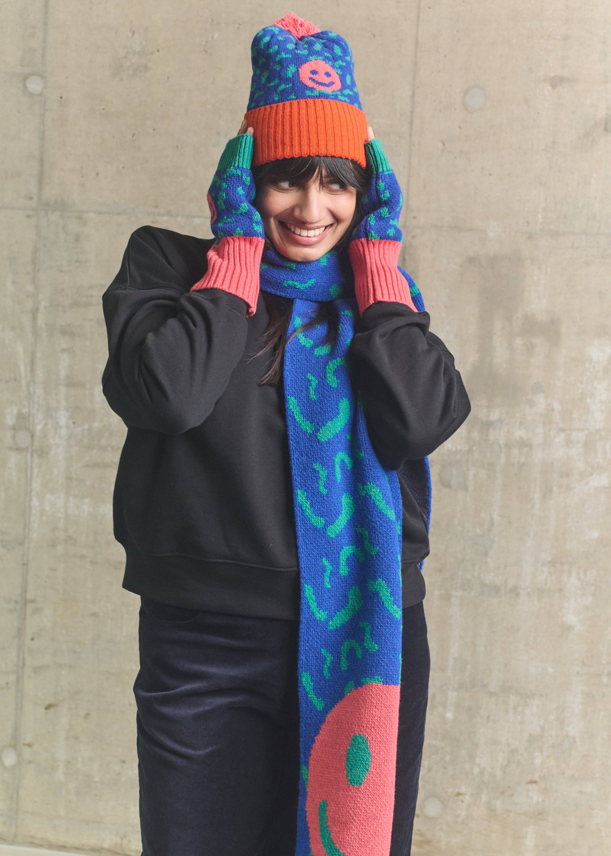 Person wearing a colorful knit hat, gloves, and scarf against a concrete wall.
