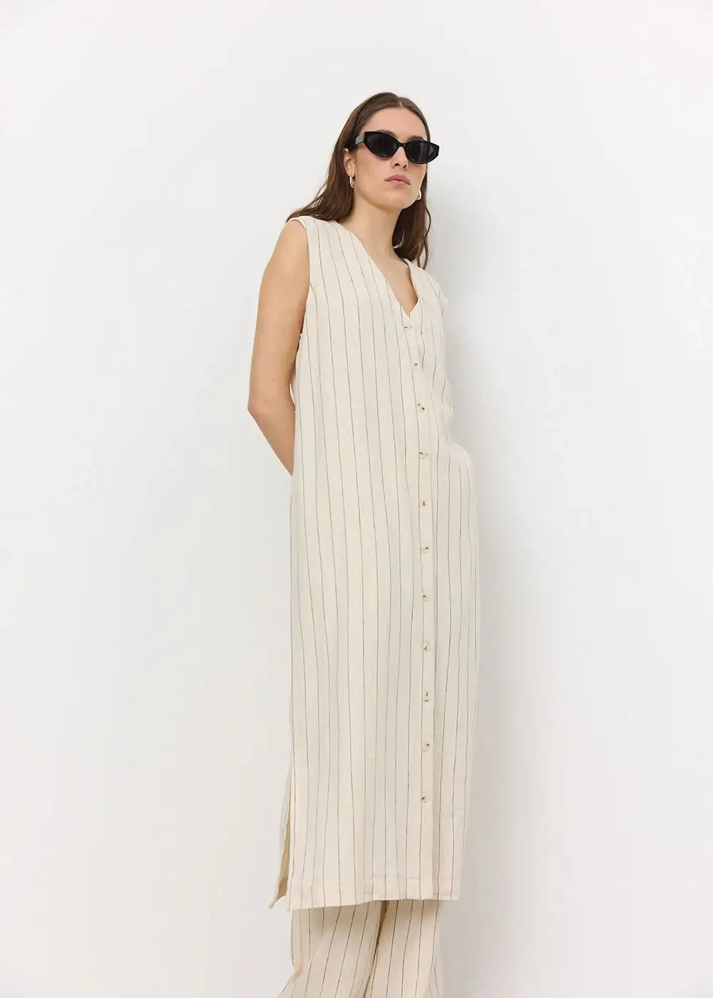 Woman wearing sleeveless cream striped button-up long dress and sunglasses against white background