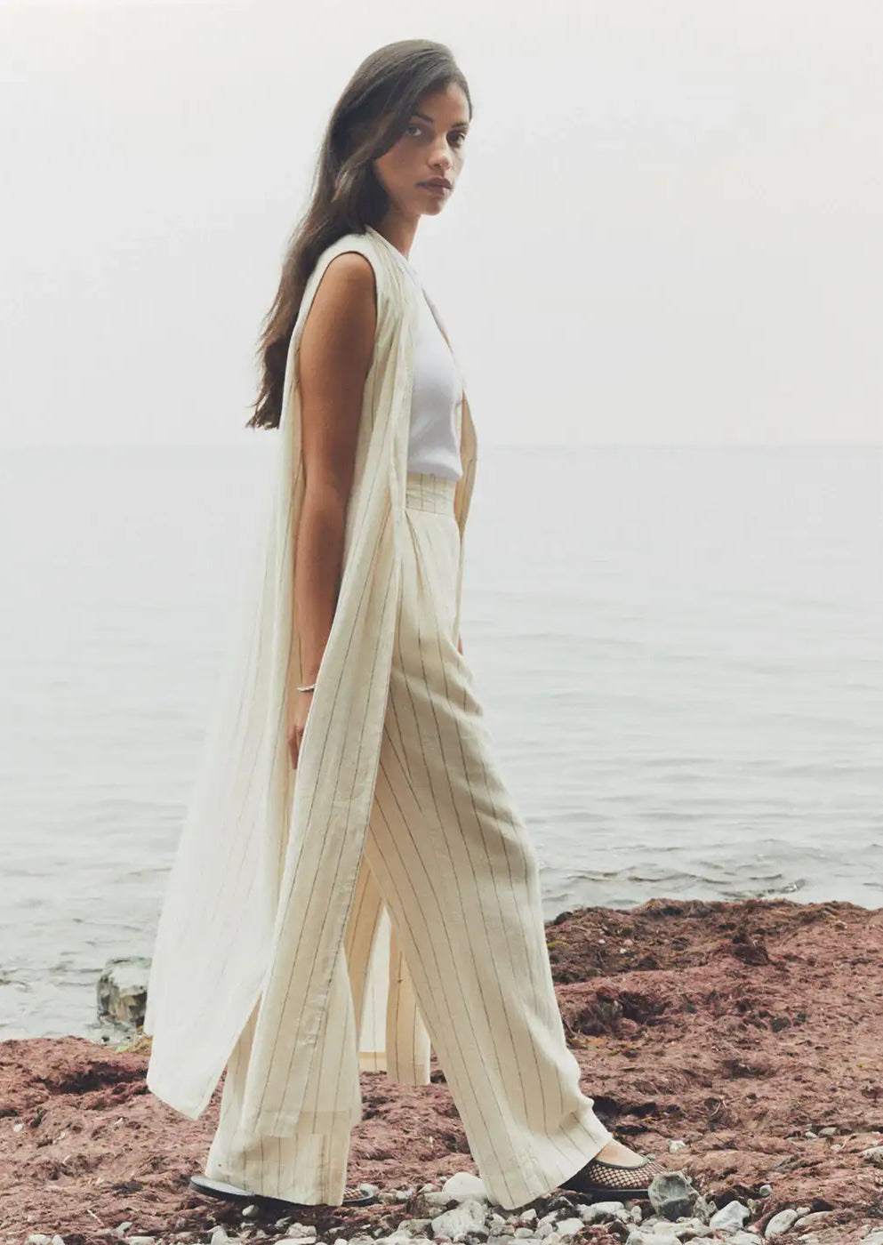 Woman in cream pinstripe sleeveless duster and wide-leg trousers by the seaside
