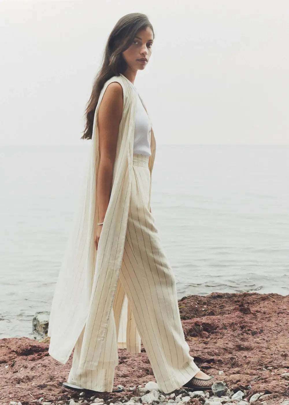 Woman in cream pinstripe sleeveless duster and wide-leg trousers by the seaside