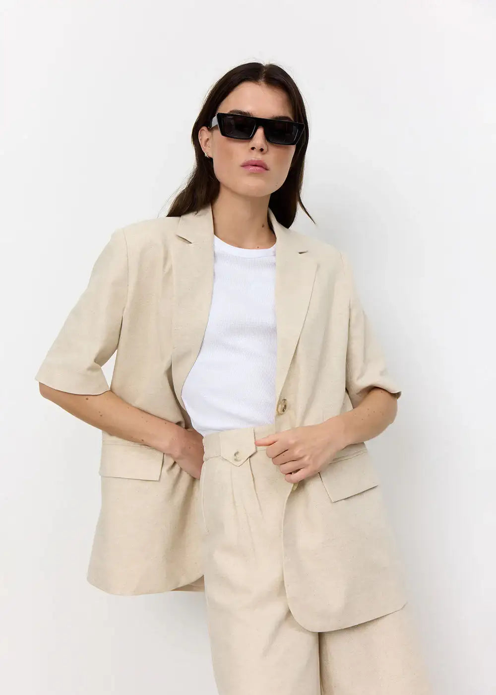 Woman in beige tailored shorts suit, white top, and black sunglasses against white background