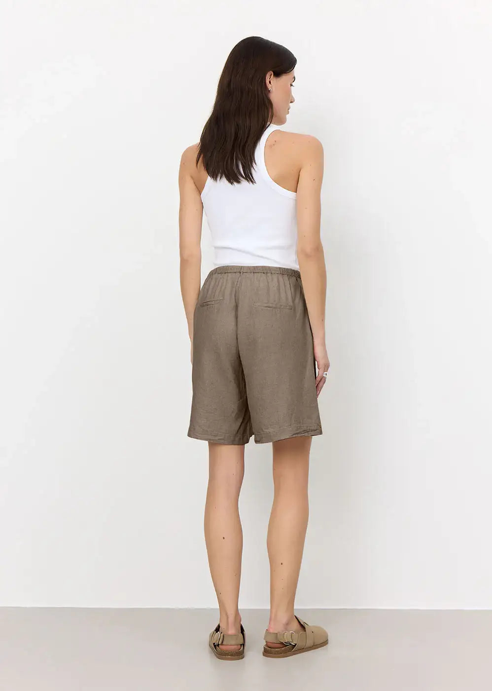 Woman in taupe linen shorts, white tank top, and sandals, seen from behind, studio setting