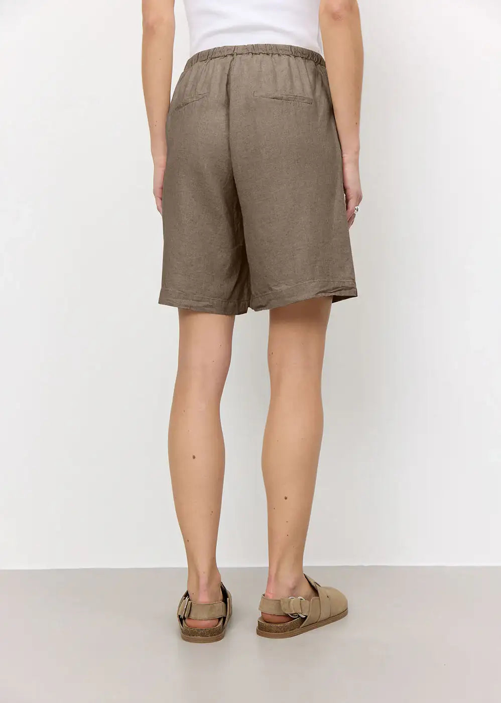 Back view of a person wearing taupe linen shorts and tan sandals against a white wall.