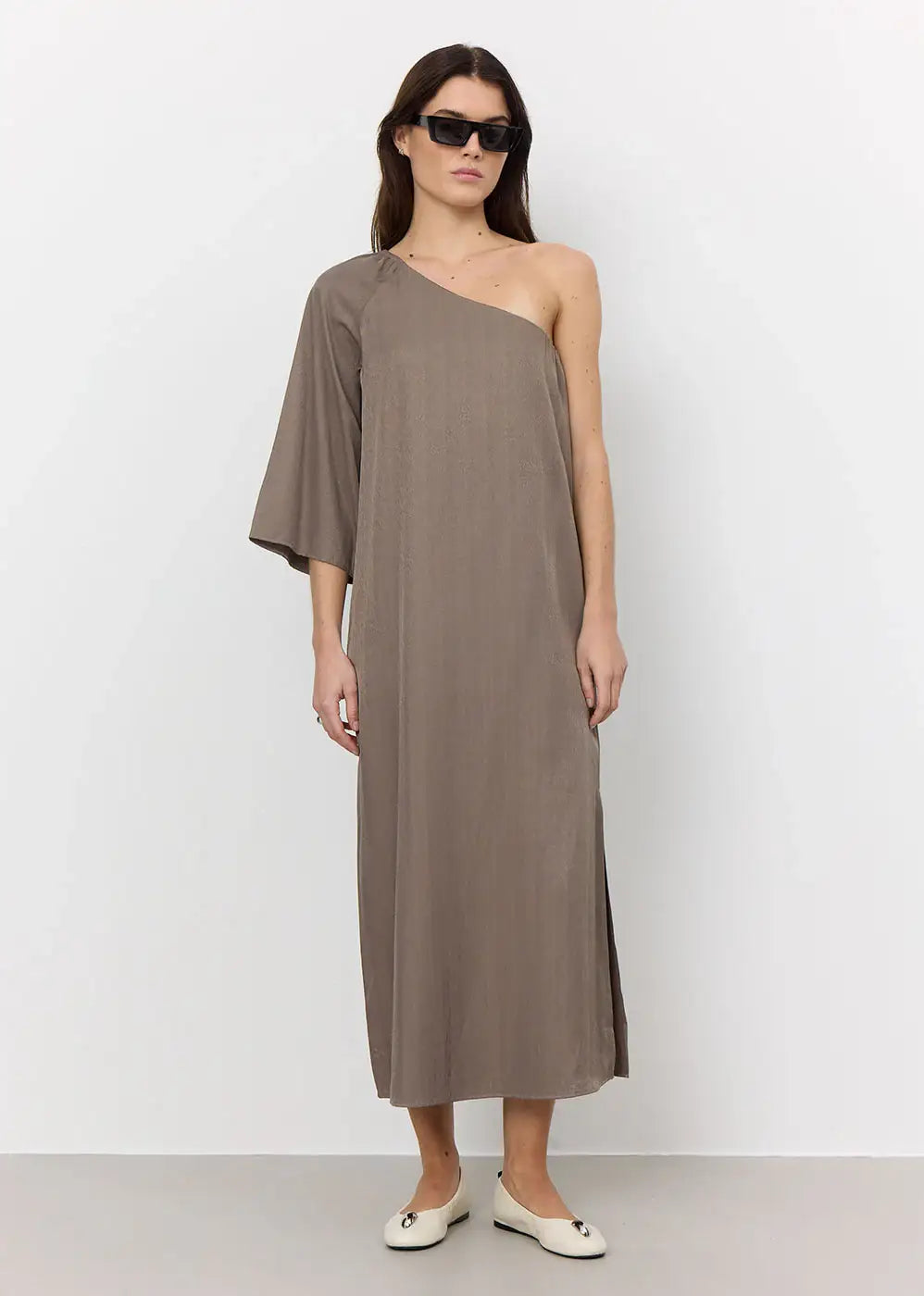 Woman wearing taupe one-shoulder maxi dress with single wide sleeve, white flats, and sunglasses