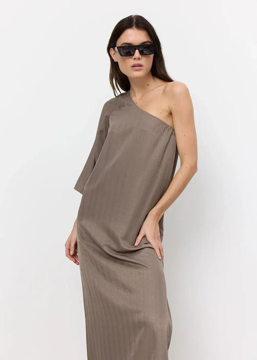 Woman wearing taupe one-shoulder dress and black sunglasses against white background