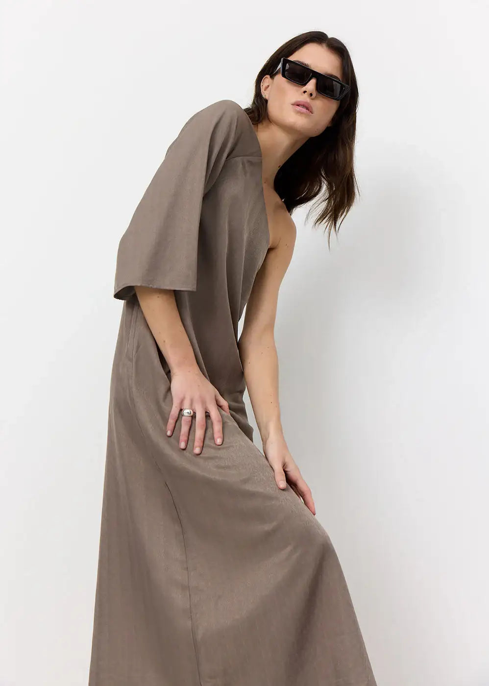 Woman in taupe asymmetric dress and sunglasses posing against white background
