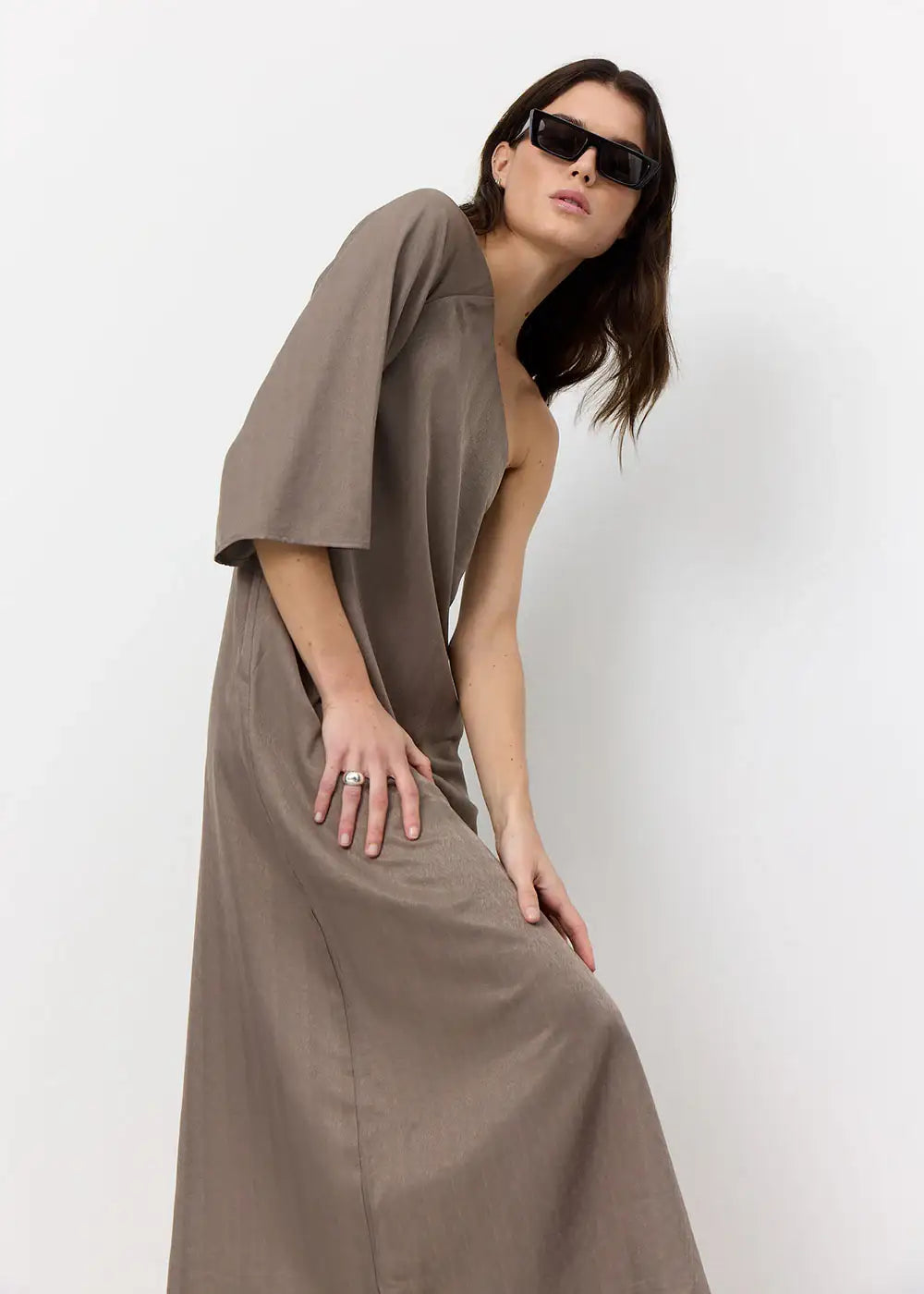 Woman in taupe asymmetric dress and sunglasses posing against white background