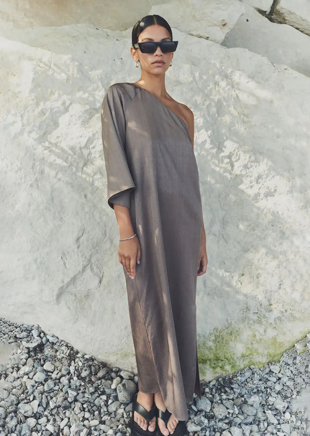 Woman in taupe one-shoulder maxi dress, sunglasses, and sandals standing on rocks outdoors