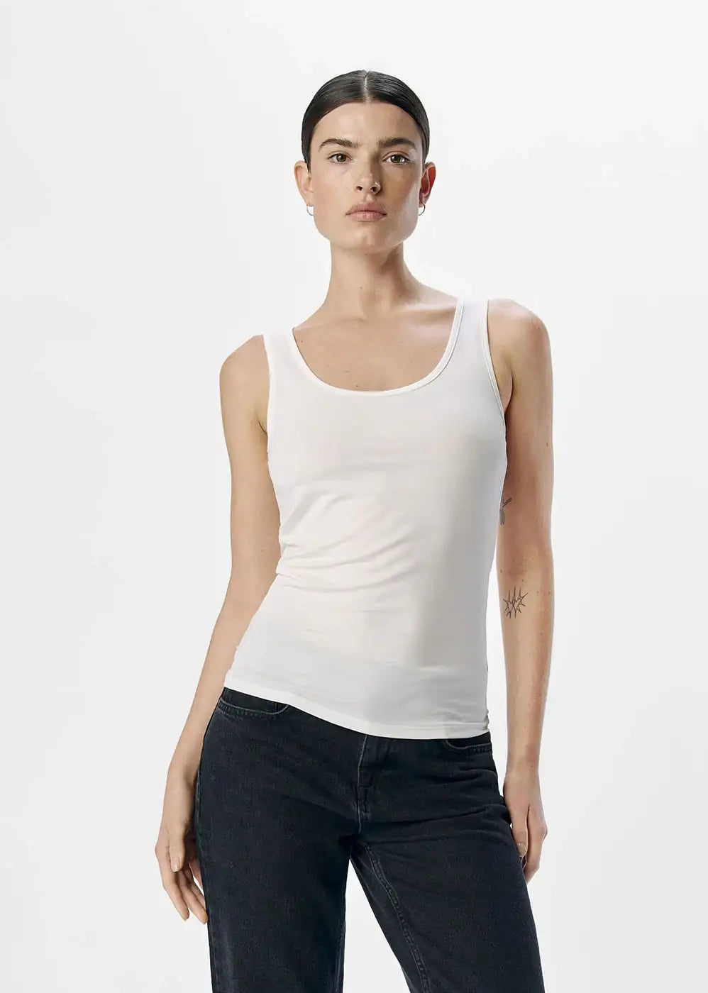 Woman wearing a fitted white tank top and black jeans on plain background