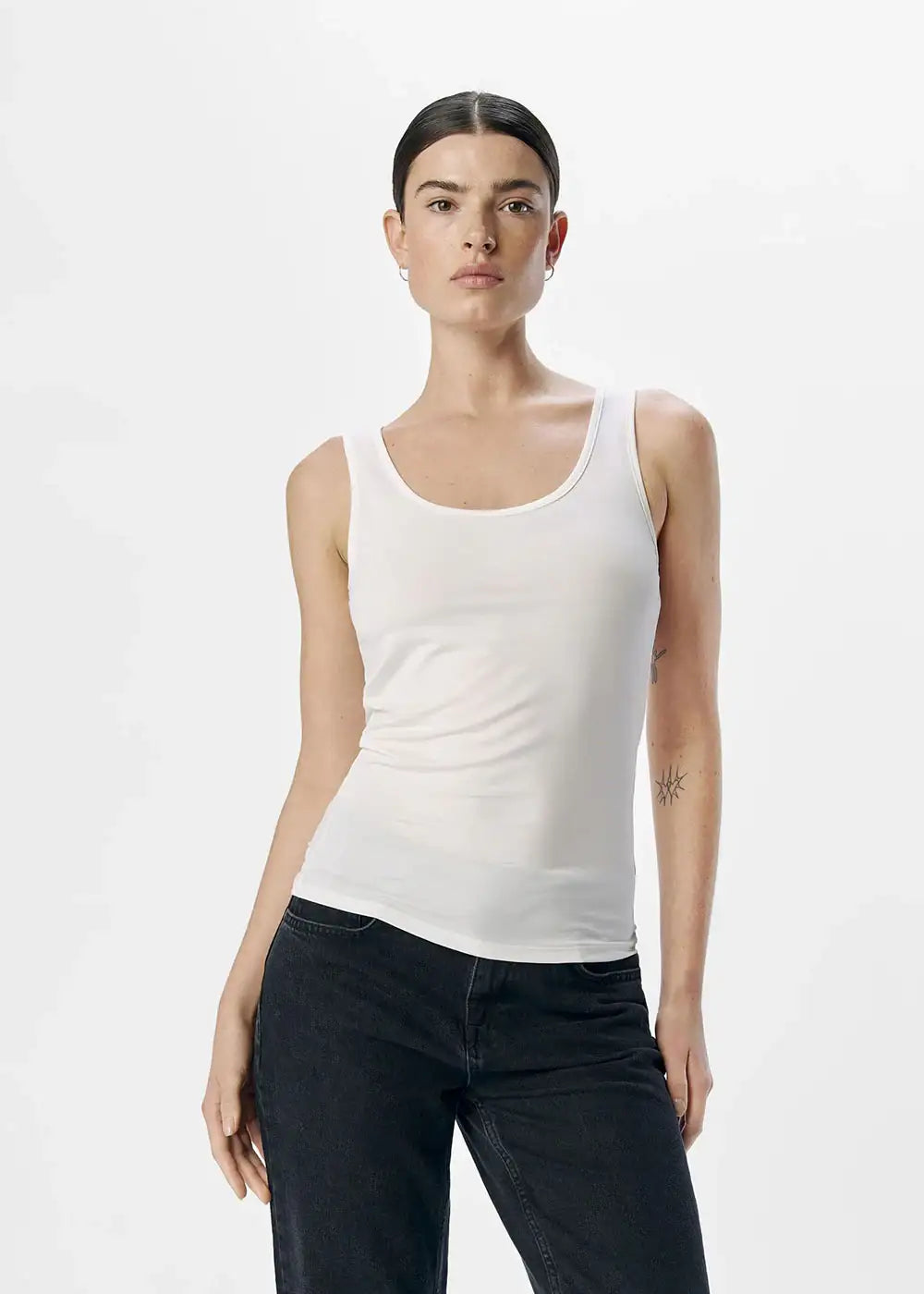 Woman wearing a fitted white tank top and black jeans on plain background