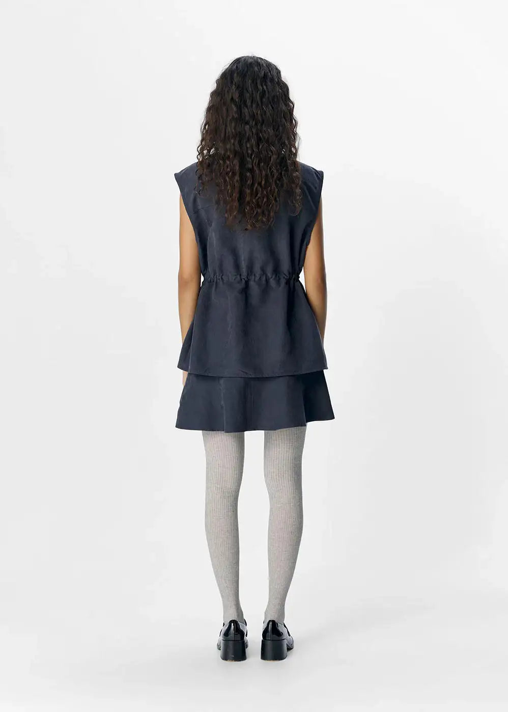 Woman wearing a dark denim sleeveless dress, grey ribbed tights, and black shoes, back view