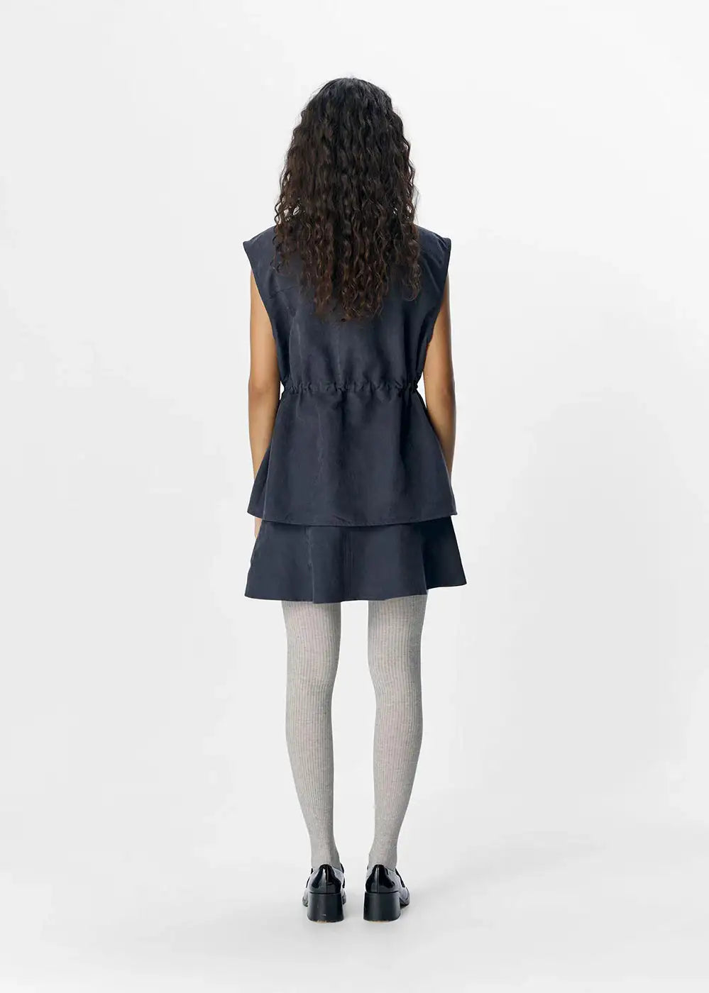 Woman wearing a dark denim sleeveless dress, grey ribbed tights, and black shoes, back view