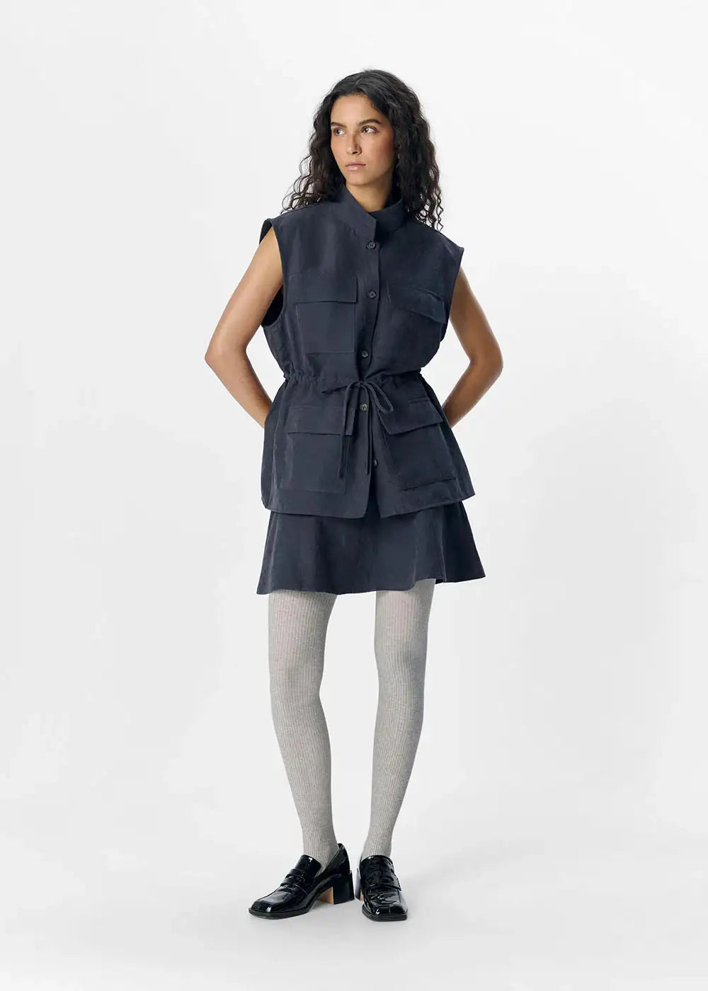 Woman in dark denim sleeveless belted vest and skirt, grey tights, black shoes, studio background