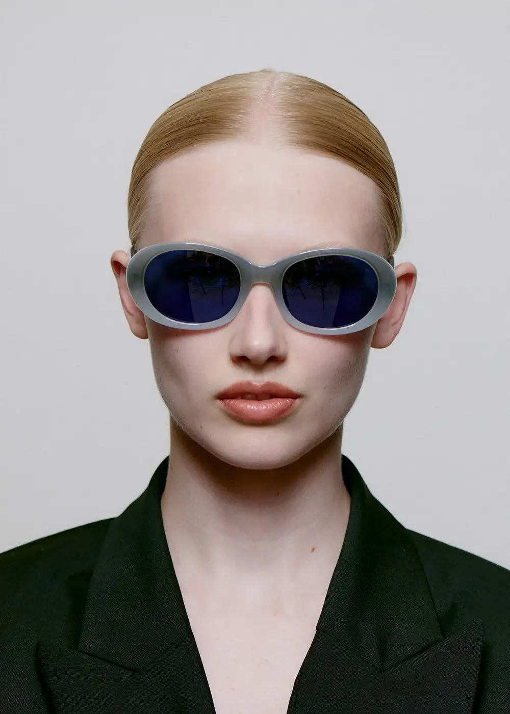 Woman wearing oval blue-lens sunglasses with grey frames, black blazer, minimalist style