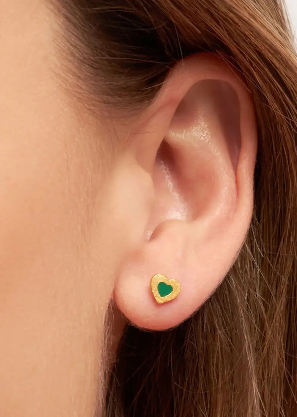 Gold plated heart-shaped green stud earring worn on ear, close-up jewellery photo