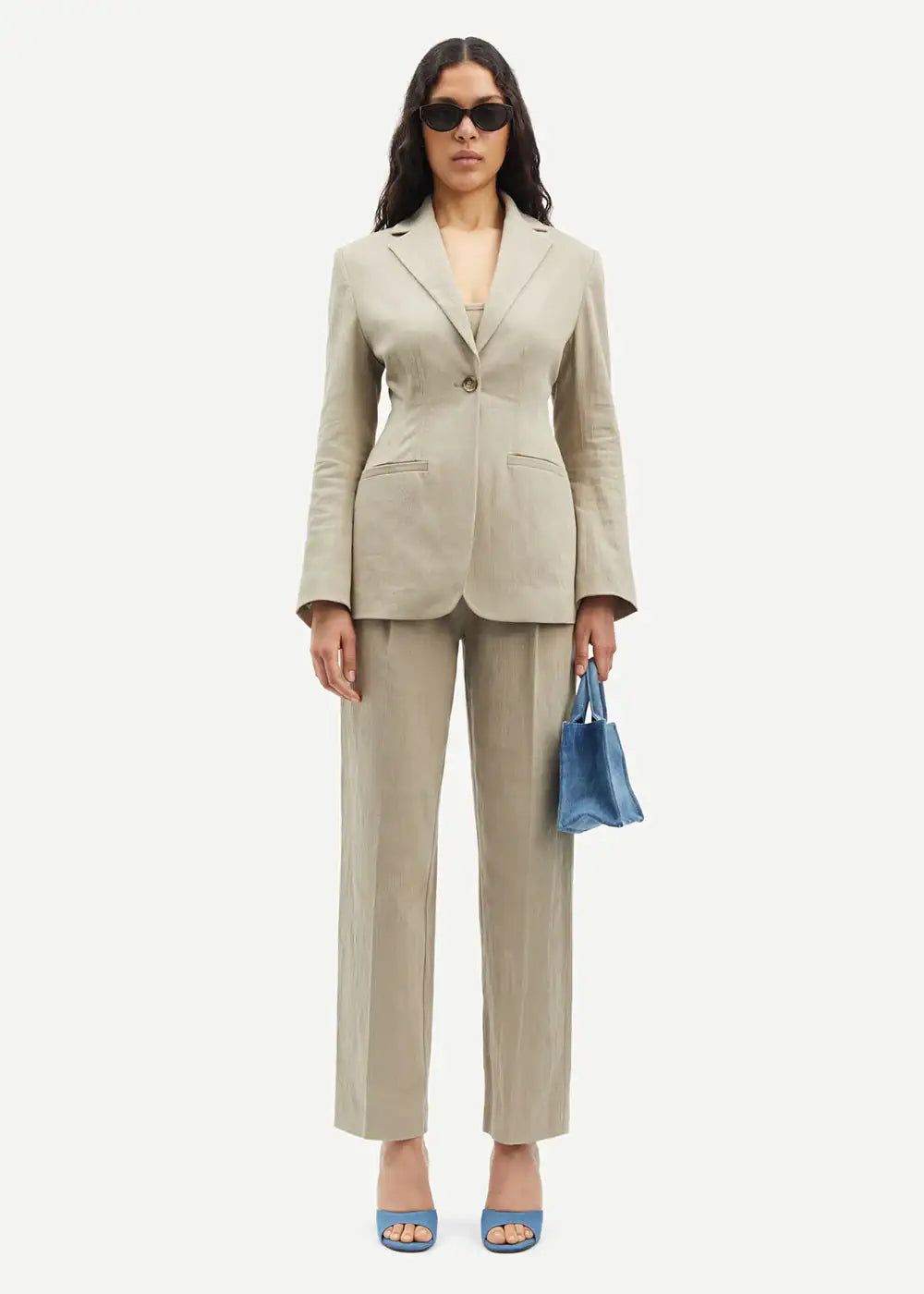 Woman wearing beige linen suit with blue heels, blue bag, and sunglasses, standing front-facing