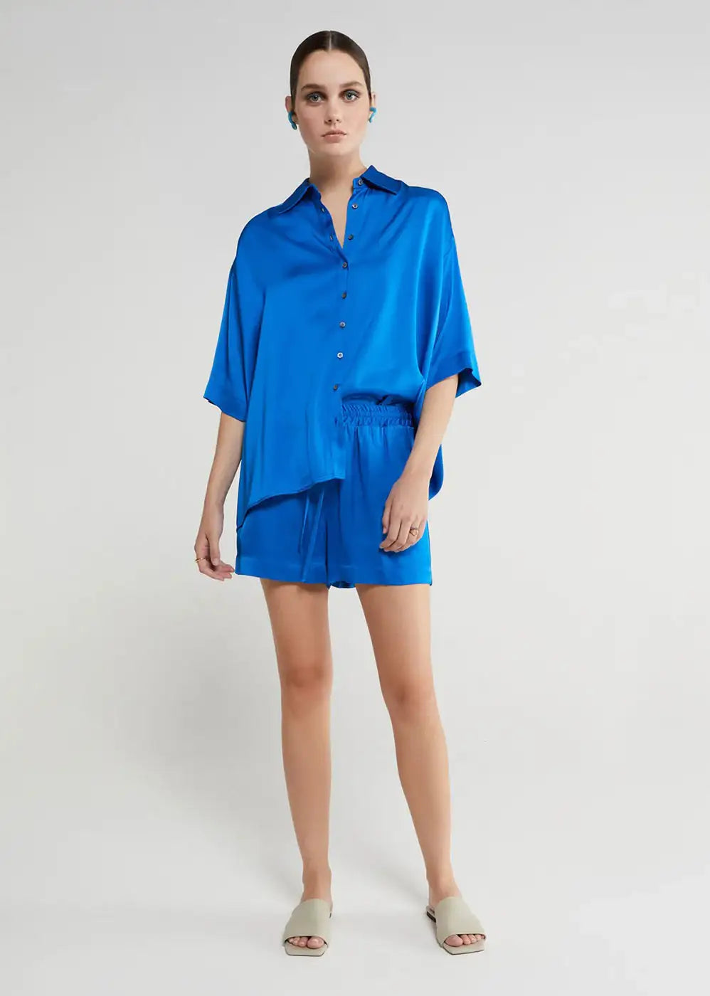 Woman in loose-fitting cobalt blue satin shirt and shorts set, standing in neutral studio