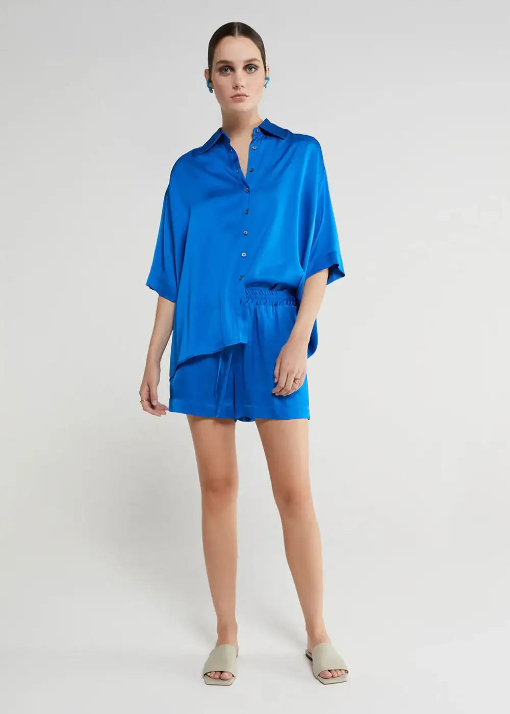 Woman in loose-fitting cobalt blue satin shirt and shorts set, standing in neutral studio
