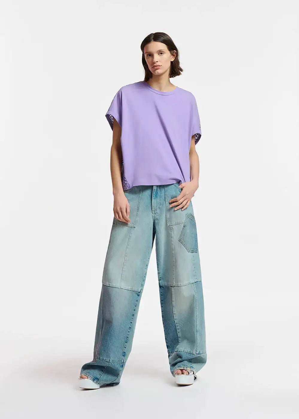 Woman wearing a loose lavender t-shirt and patchwork wide-leg denim jeans on white background