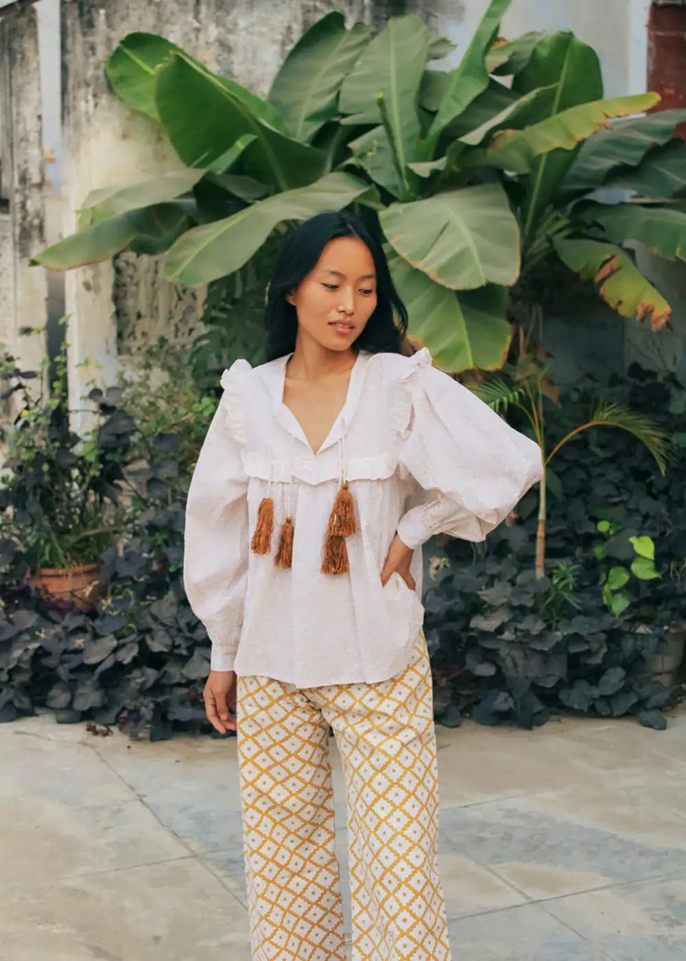 Woman in white blouse with tassels and patterned wide-leg pants, standing outdoors with tropical plants