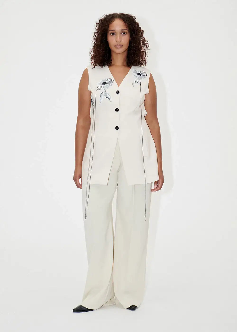 Woman wearing ivory wide-leg pants and sleeveless embroidered vest in minimalist setting
