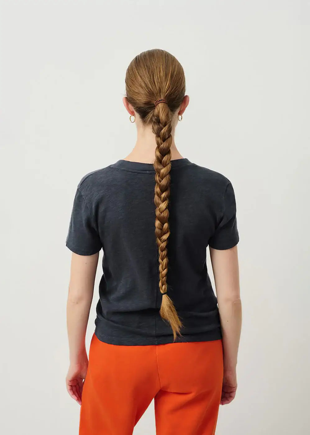 Woman with long braided hair wearing a dark t-shirt and orange trousers, back view