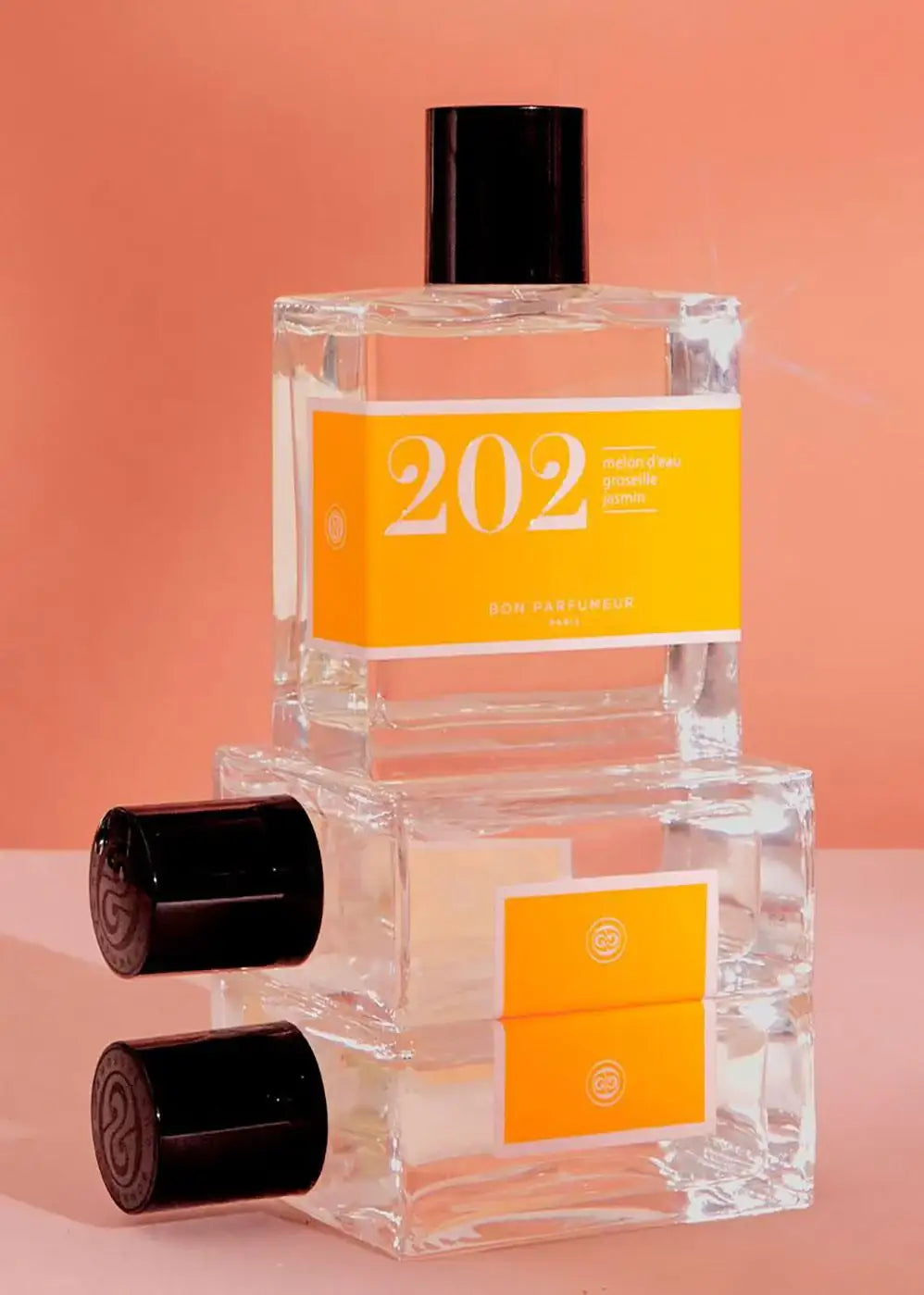 Stacked clear glass perfume bottles with orange labels and black caps on a peach background