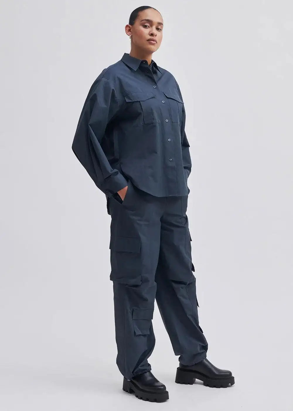 Woman in navy oversized shirt and cargo pants set with chunky black boots, studio background