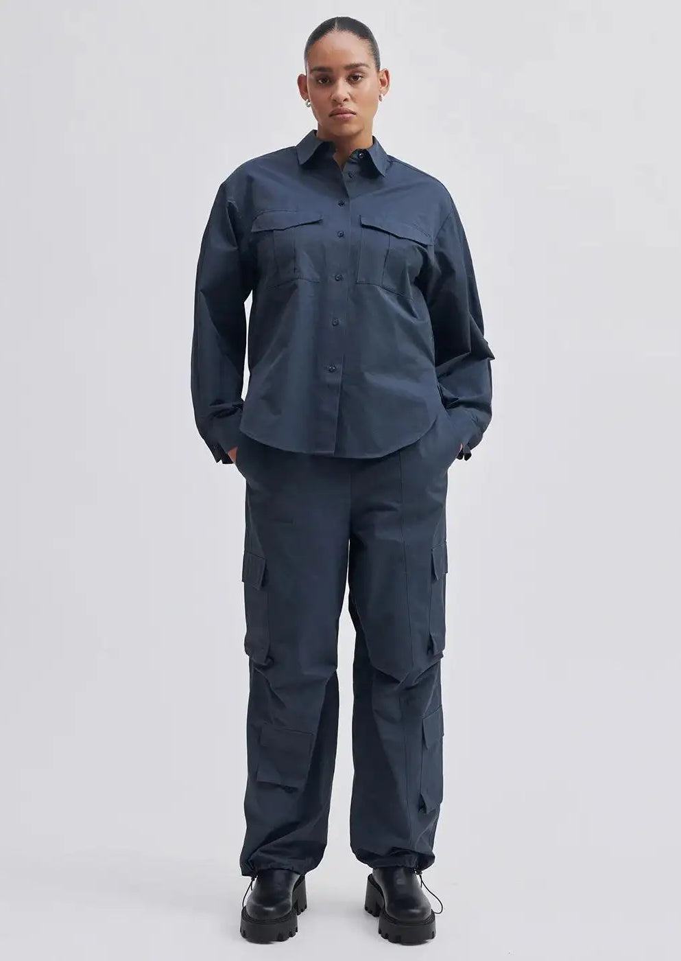 Woman wearing dark navy cargo trousers and matching button-up shirt, black boots, neutral background