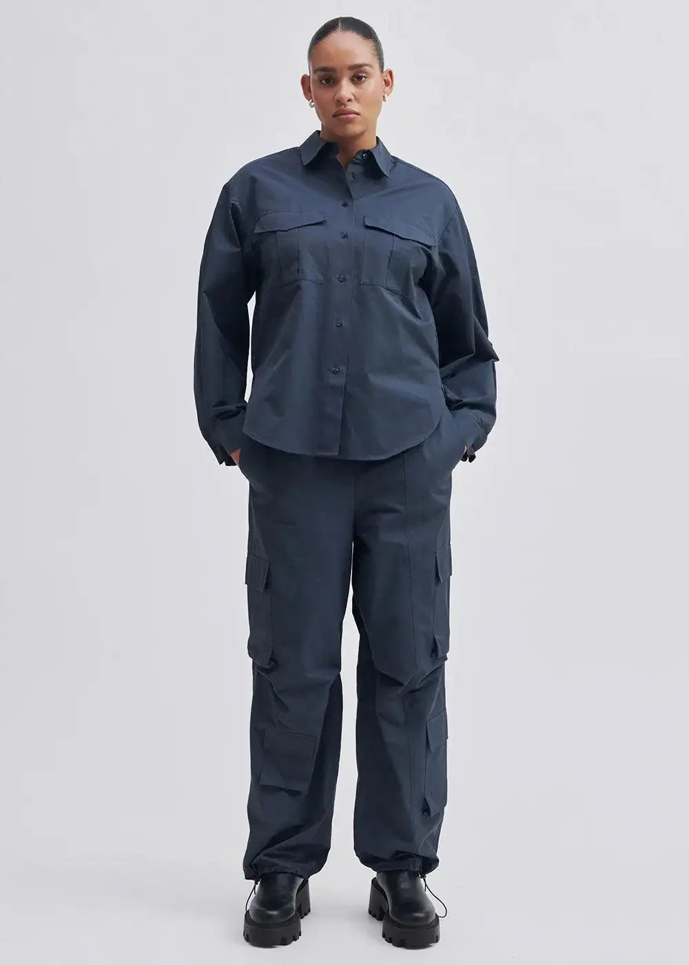 Woman wearing dark navy cargo trousers and matching button-up shirt, black boots, neutral background