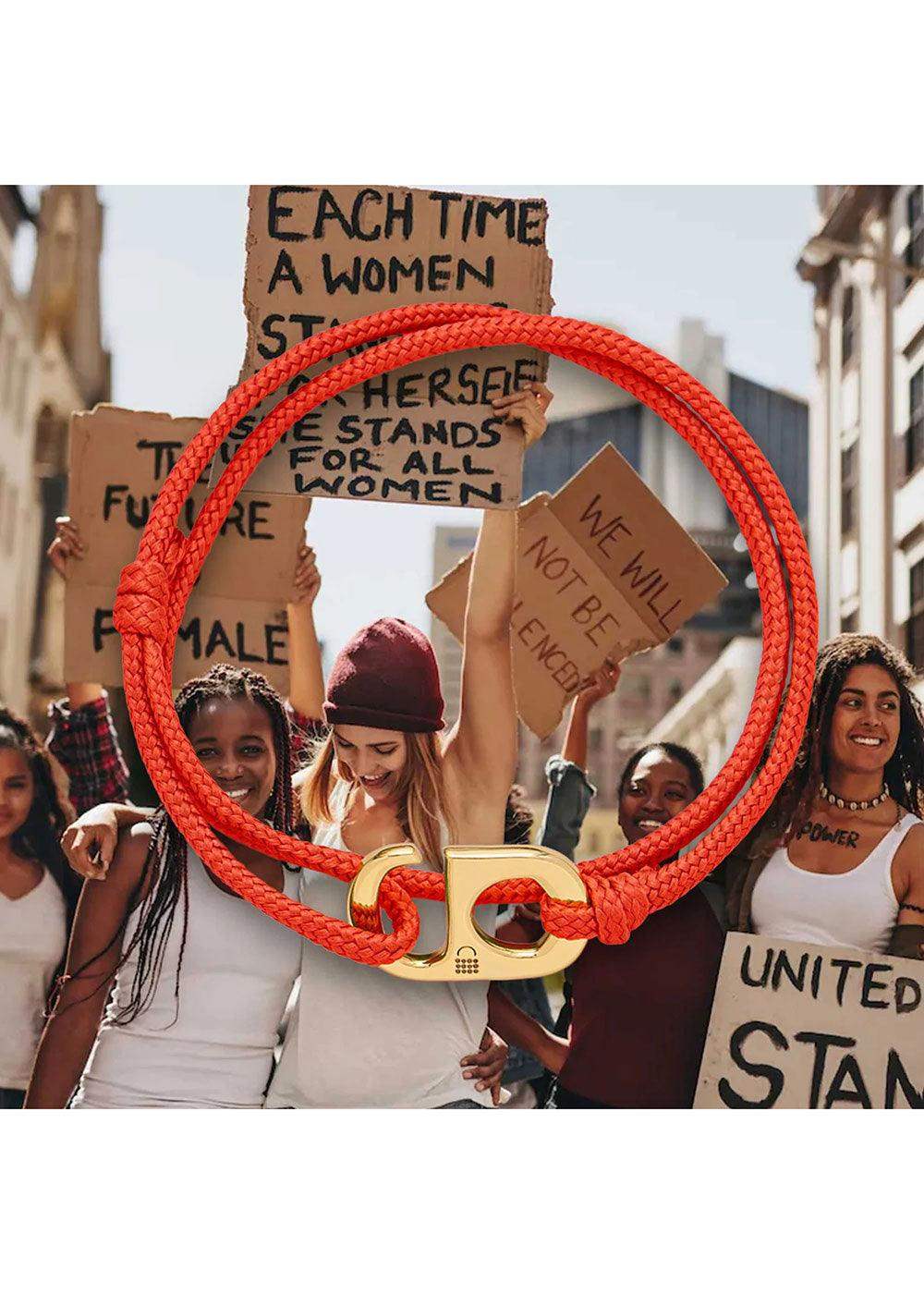 Red cord bracelet with gold clasp over women at equality rally holding protest signs