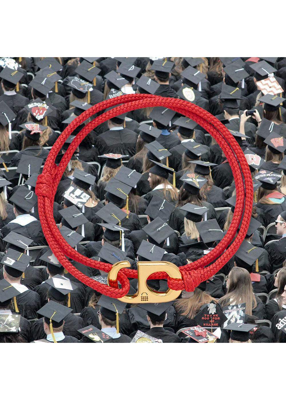 Red braided bracelet with gold clasp displayed over a crowd of graduates in black caps and gowns