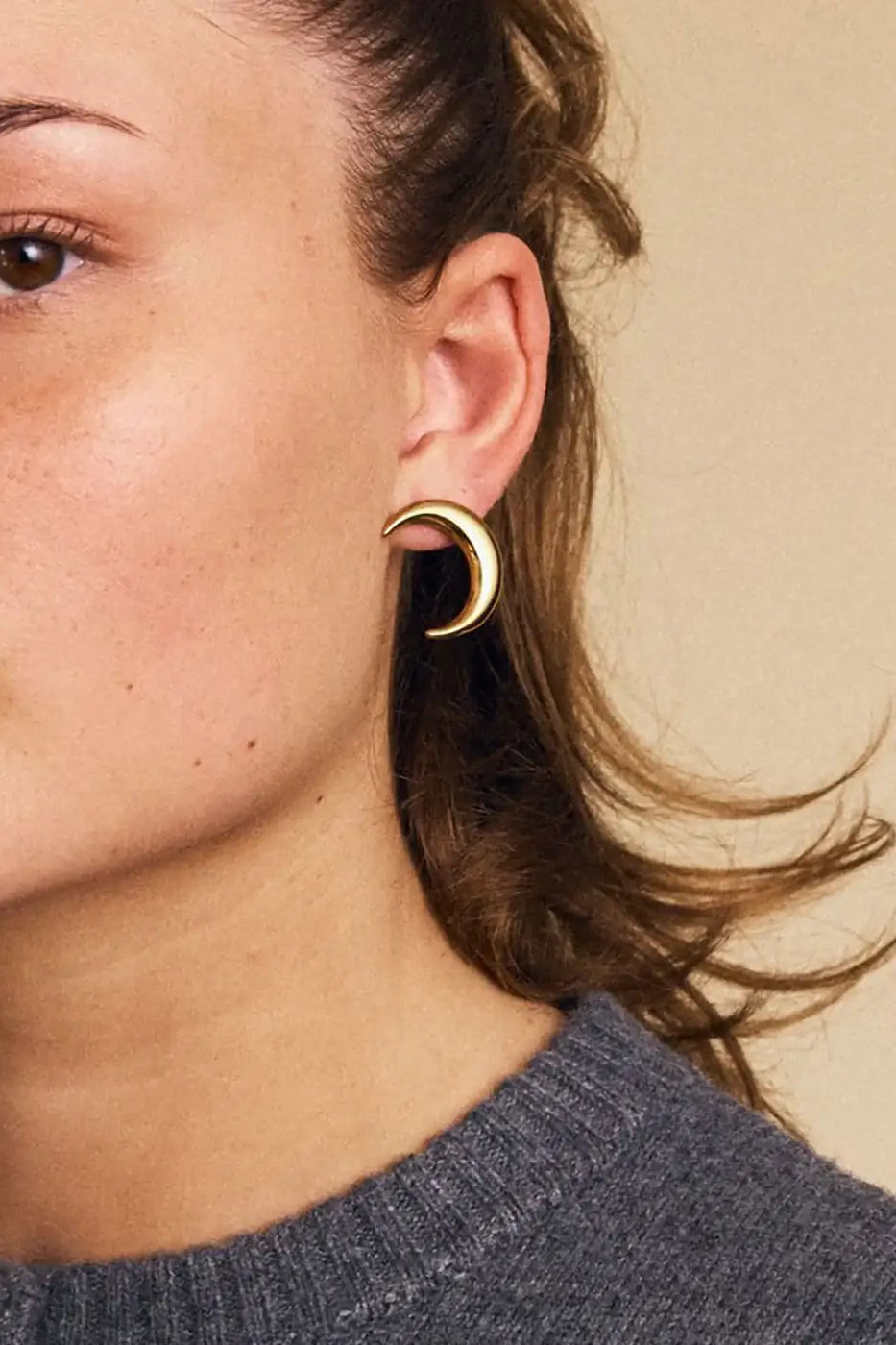 Gold crescent moon stud earring worn on ear, modern jewelry against neutral background