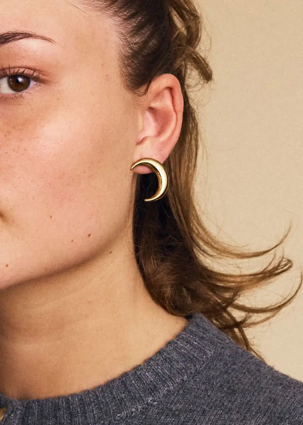 Gold crescent moon stud earring worn on ear, modern jewelry against neutral background