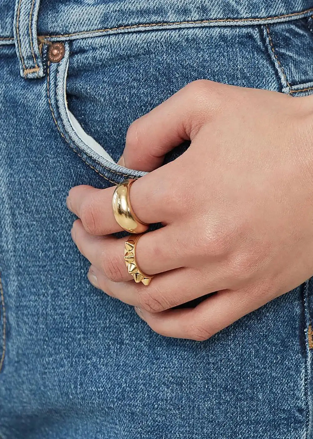 Hand wearing chunky gold rings with studded detail, resting on blue denim jeans