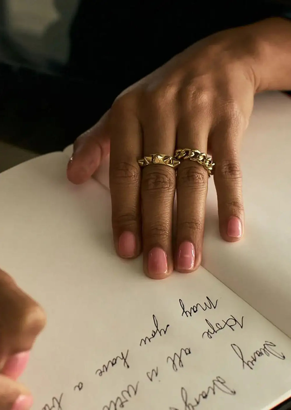 Hand with two chunky gold rings writing in a notebook, polished pink nails visible