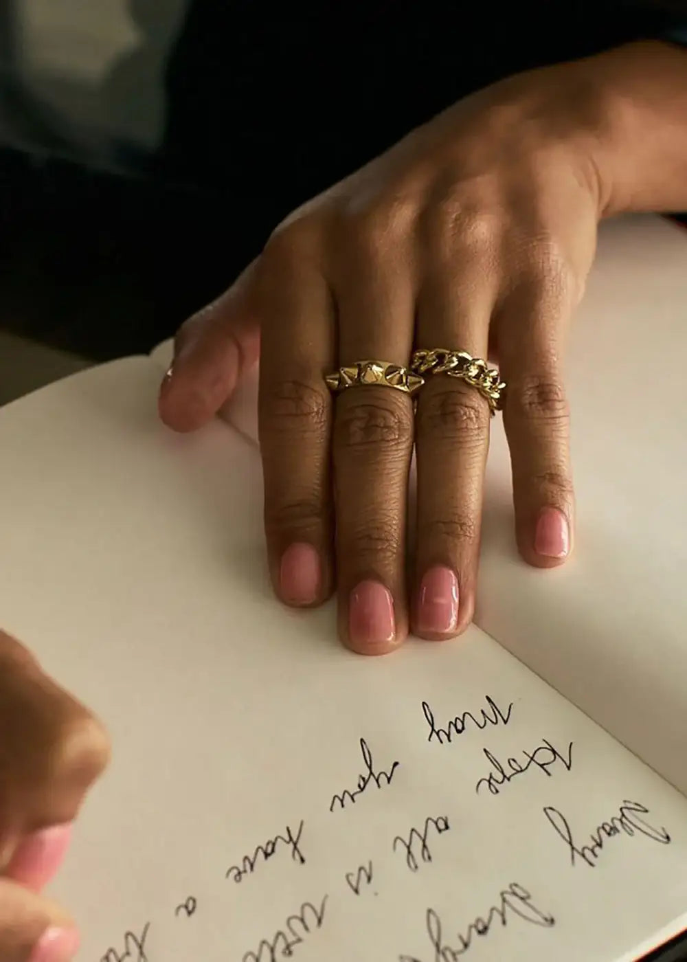 Hand with two chunky gold rings writing in a notebook, polished pink nails visible