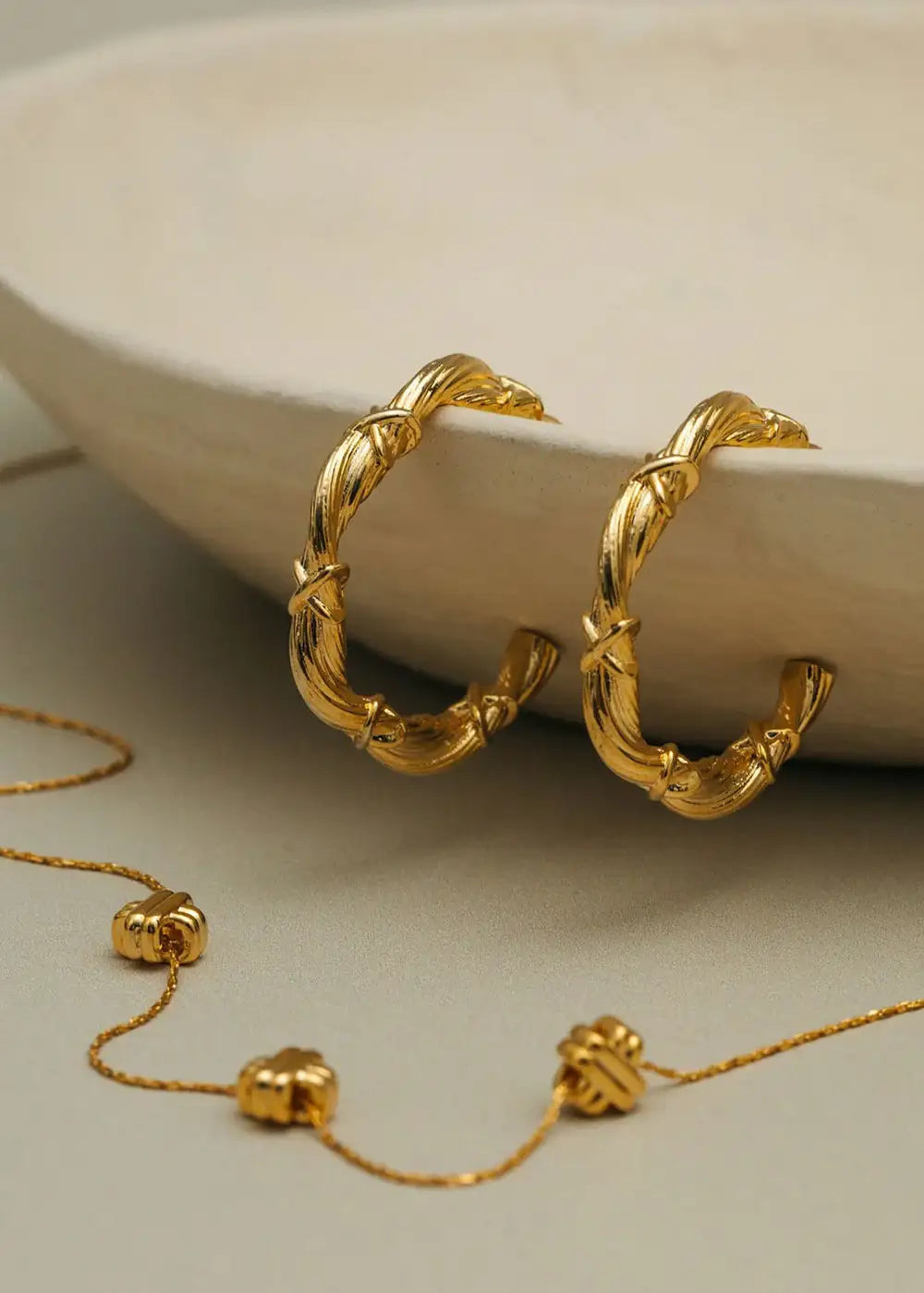 Gold twist and cross hoop earrings displayed on a neutral dish with gold chain accents