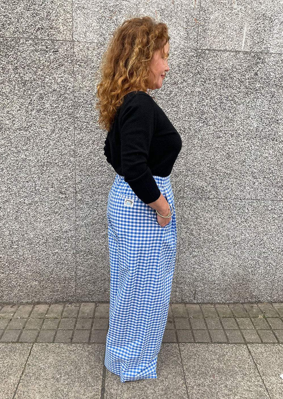 Woman in black top and blue gingham wide-leg trousers standing by grey stone wall