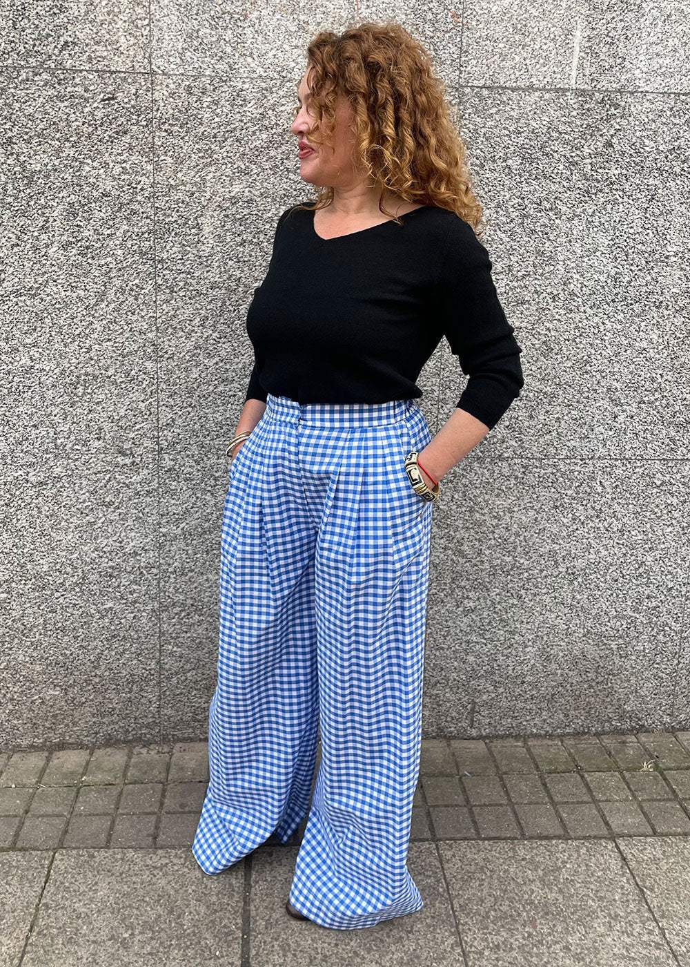 Woman wearing black top and wide-leg blue gingham trousers standing against stone wall