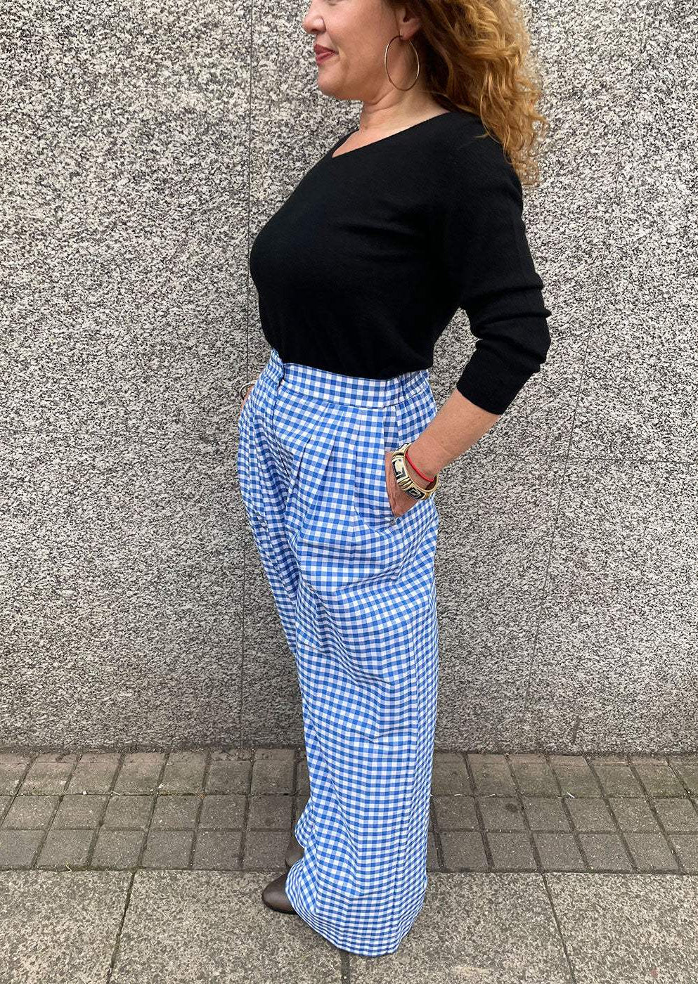 Woman wearing blue gingham wide-leg trousers and black top, standing by stone wall