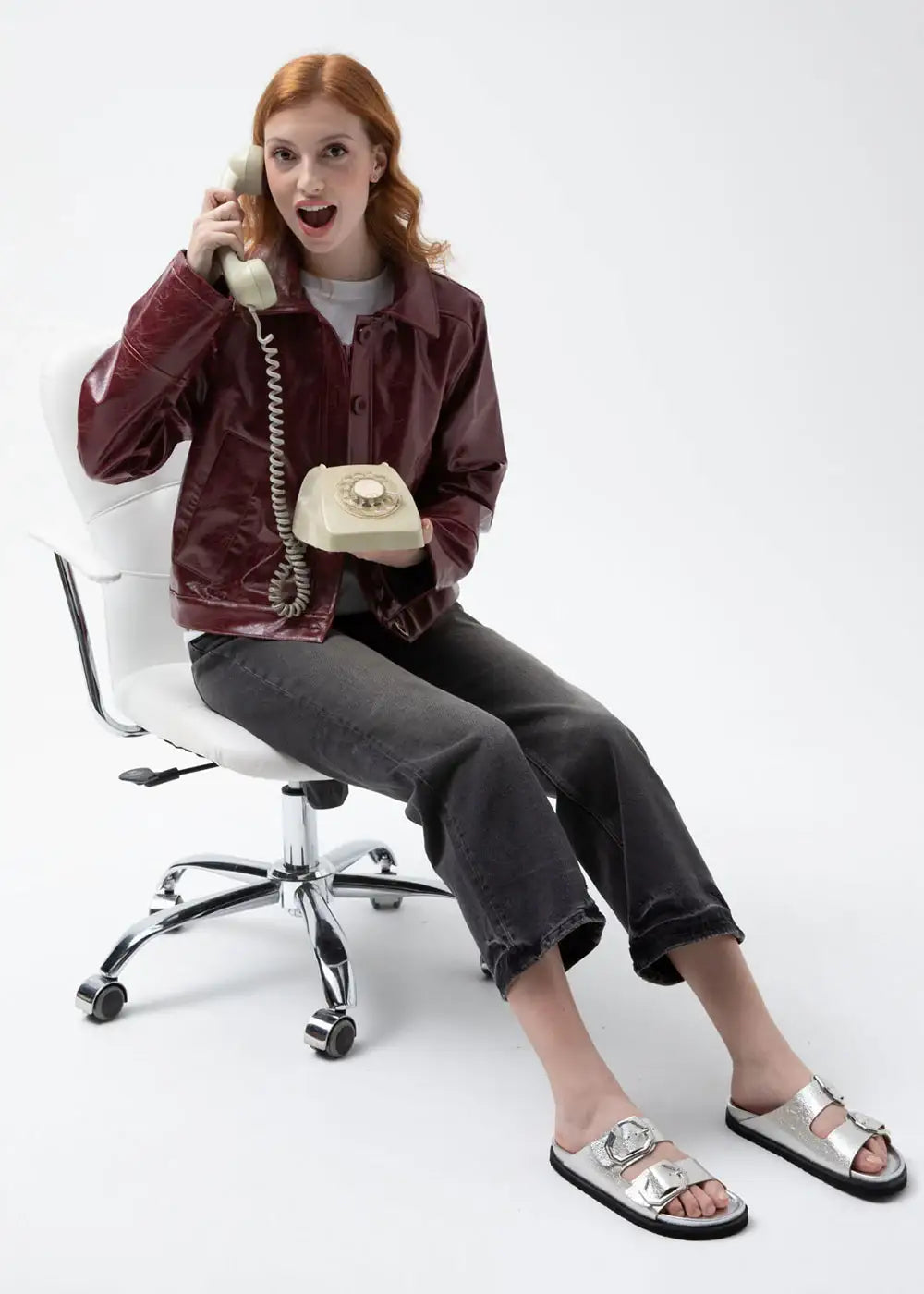 Woman in a burgundy leather jacket, cropped jeans, and silver sandals on a white chair holding a vintage rotary phone