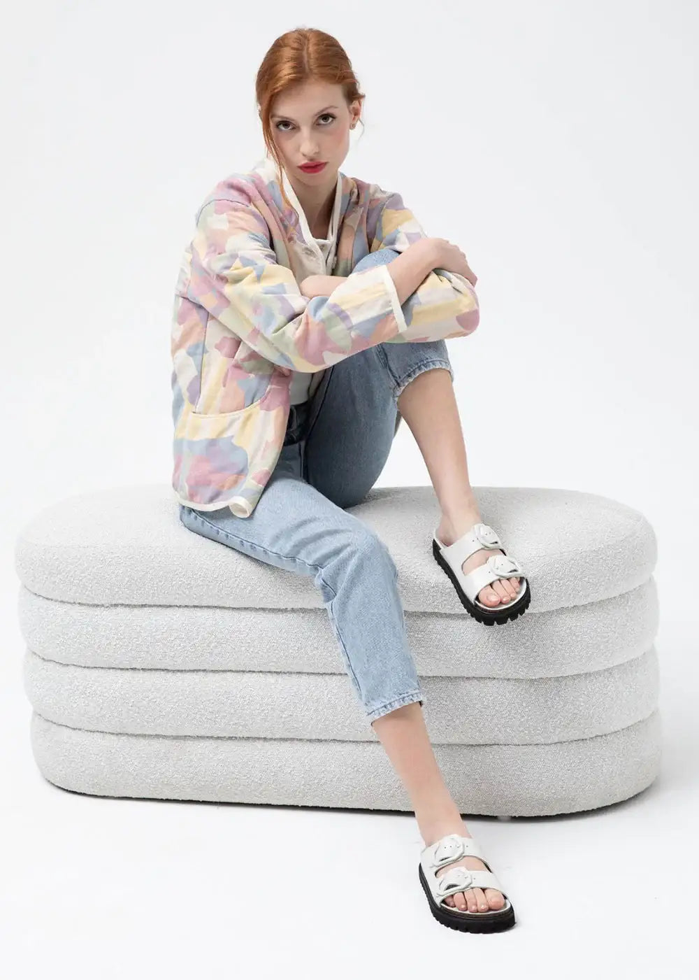 Woman in pastel patchwork jacket, light jeans, and white sandals sitting on textured bench