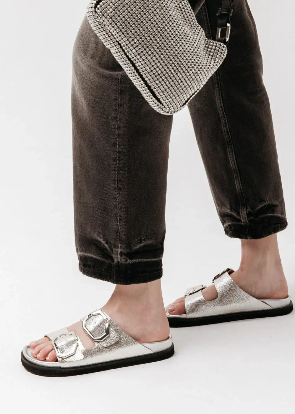 Woman wearing silver buckle sandals, black jeans, and a silver mesh purse