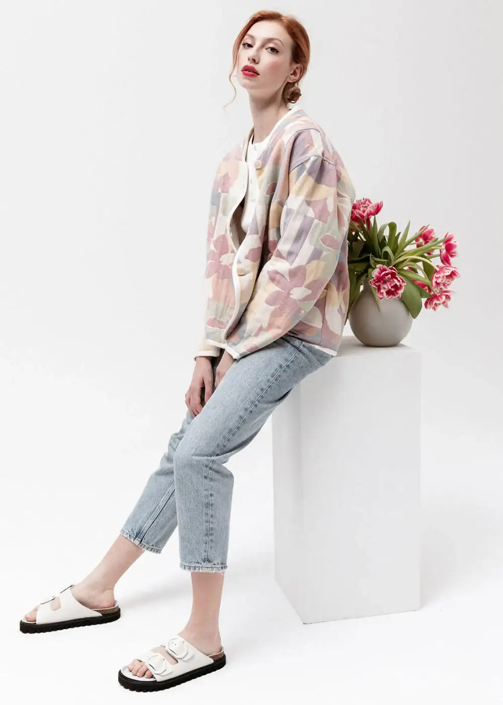 Woman in pastel floral jacket, light jeans, and white sandals seated by vase of pink tulips