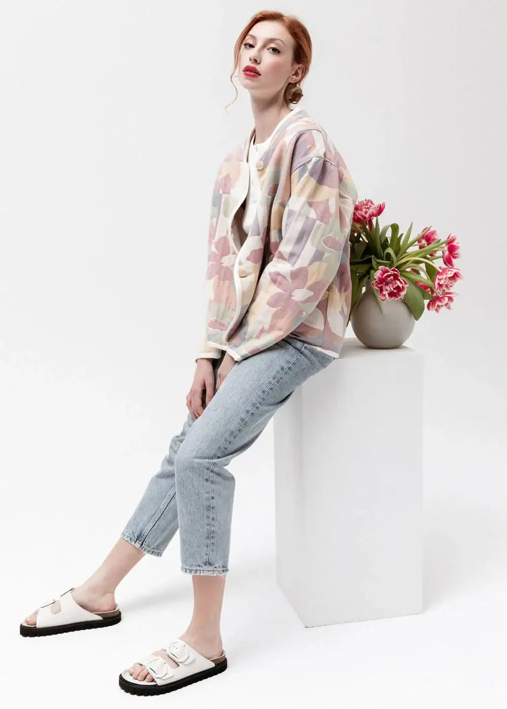 Woman in pastel floral jacket, light jeans, and white sandals seated by vase of pink tulips