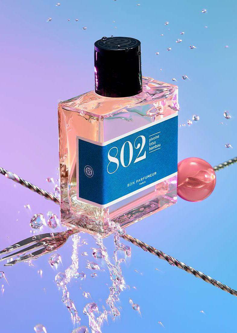 Clear glass perfume bottle labeled 802 with blue label, water splashes, and a pink sphere on a pastel background.