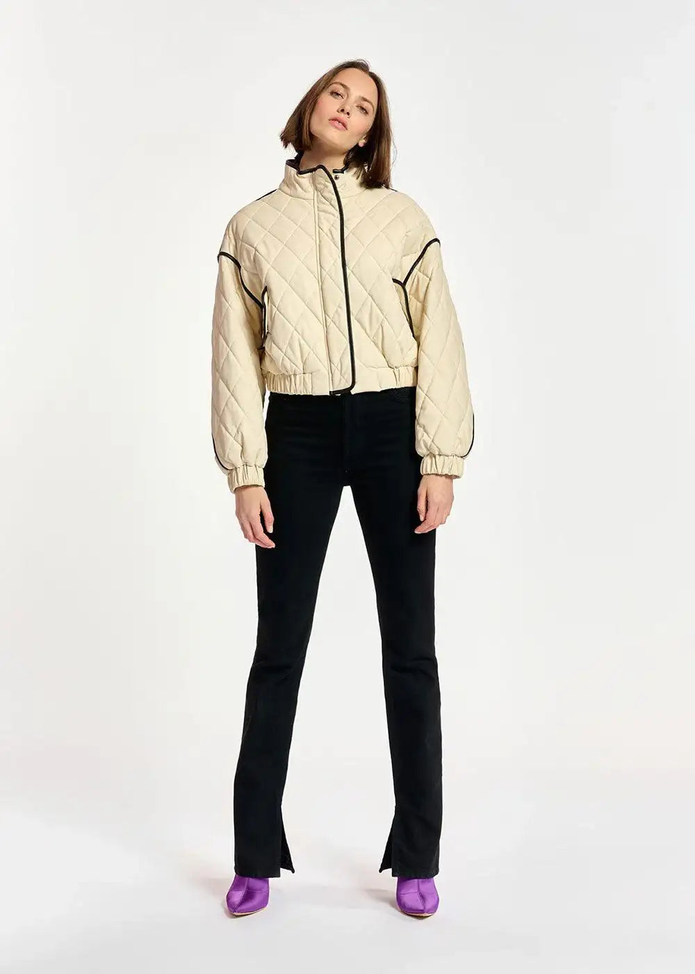 Woman wearing black split hem jeans, cream quilted jacket, and purple boots on white background.