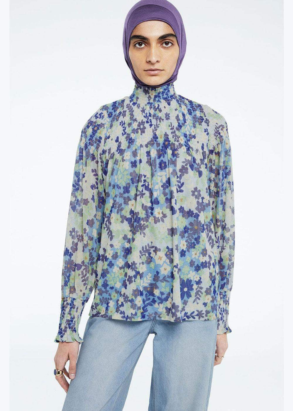 Woman in blue floral long sleeve blouse with high neck, light wash jeans, and purple headscarf