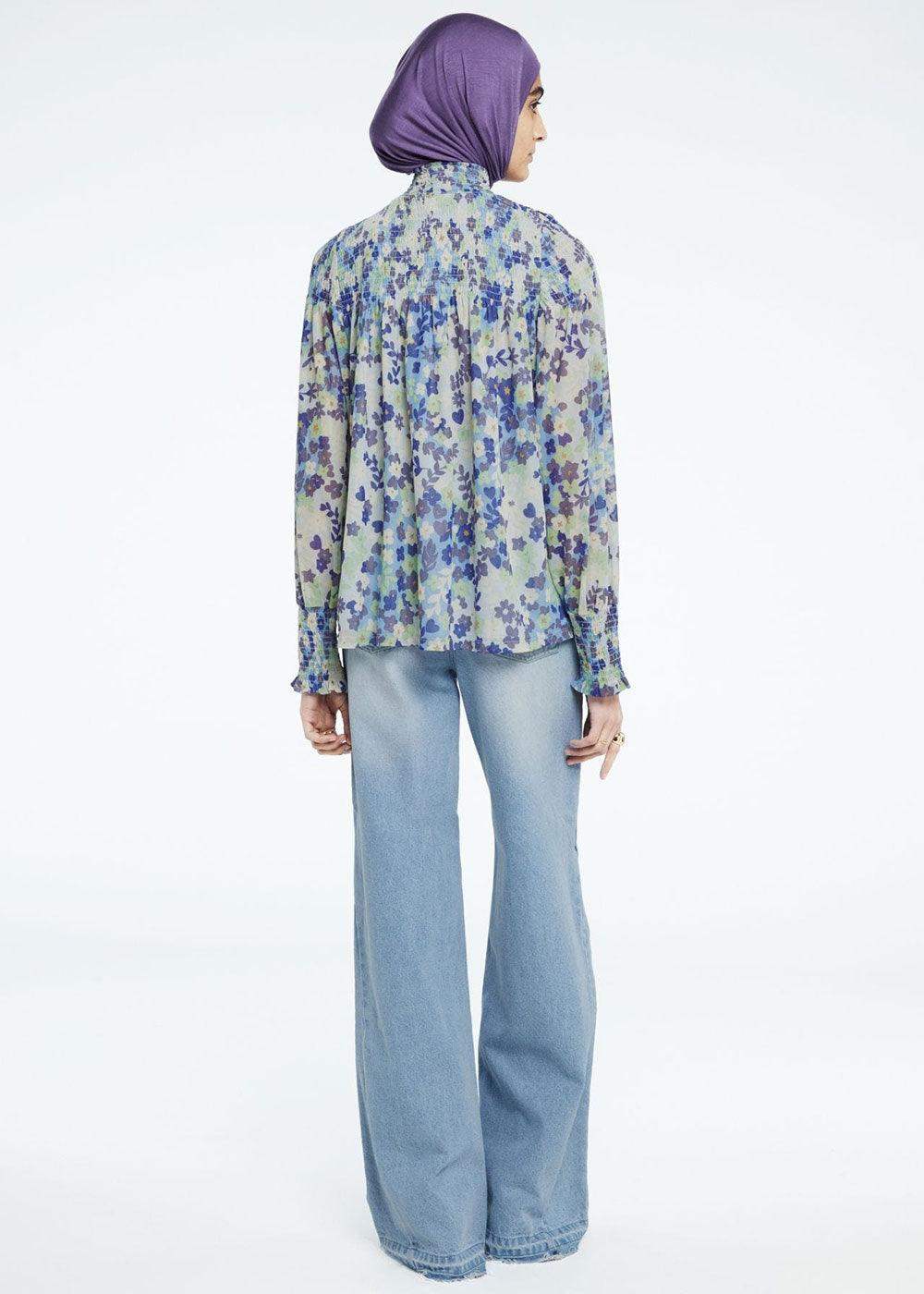 Woman in purple headscarf, floral sheer blouse, and light blue flared jeans, back view